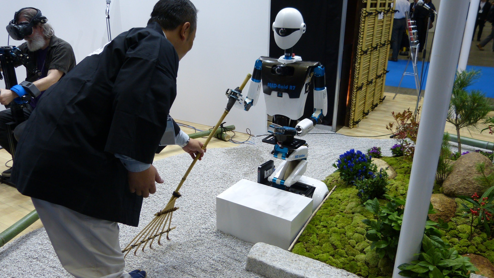 a japanese man hands a robot a rake near some sand and plants.  Earl is in the background using the vr system.